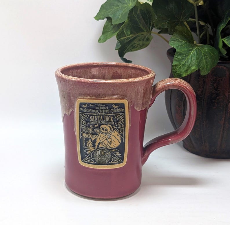 May include: A ceramic mug with a glossy cranberry red glaze and a lighter drip effect at the rim. A rectangular label features a black and cream graphic of Santa Jack from The Nightmare Before Christmas, including text and a spiderweb design.