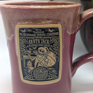 May include: A cranberry-colored ceramic mug with a reactive glaze rim. A rectangular plaque features a raised black and tan graphic of Santa Jack from The Nightmare Before Christmas, with text reading Santa Jack, Cranberry Creme Brulee, and Bones.