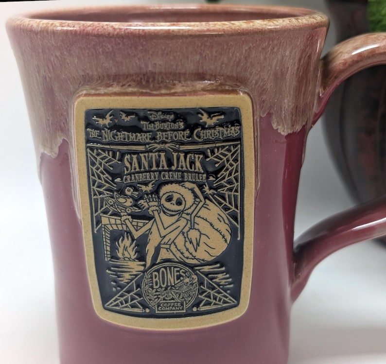 May include: A cranberry-colored ceramic mug with a reactive glaze rim. A rectangular plaque features a raised black and tan graphic of Santa Jack from The Nightmare Before Christmas, with text reading Santa Jack, Cranberry Creme Brulee, and Bones.