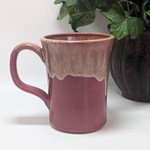 May include: A ceramic mug featuring a deep rose-colored base and a contrasting cream-colored glaze that drips down from the rim. The mug has a sturdy, curved handle on the left side and a smooth, glossy finish throughout its cylindrical body.