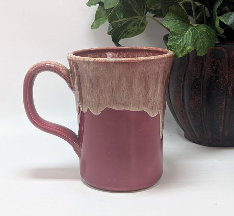 May include: A ceramic mug featuring a deep rose-colored base and a contrasting cream-colored glaze that drips down from the rim. The mug has a sturdy, curved handle on the left side and a smooth, glossy finish throughout its cylindrical body.