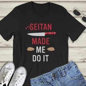 May include: Black t-shirt with white text that reads "Seitan Made Me Do It". The text is surrounded by a red knife and two brown seitan patties.