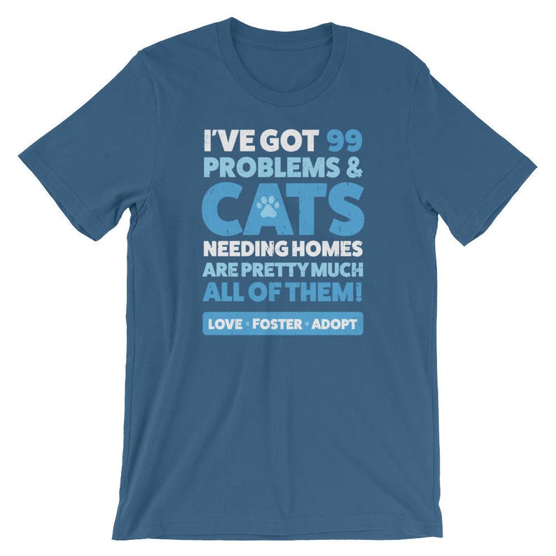 Puede incluir: Una camiseta azul con un gr&aacute;fico blanco que dice "Tengo 99 problemas y gatos que necesitan hogar, casi todos! Amor, acogida, adopci&oacute;n."