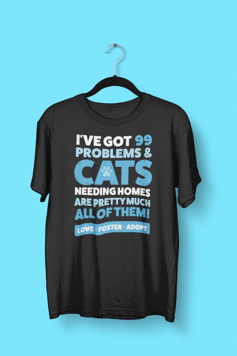 Puede incluir: Camiseta negra con un gr&aacute;fico azul que dice "Tengo 99 problemas y gatos que necesitan hogar, &iexcl;son casi todos! Amar, acoger, adoptar."