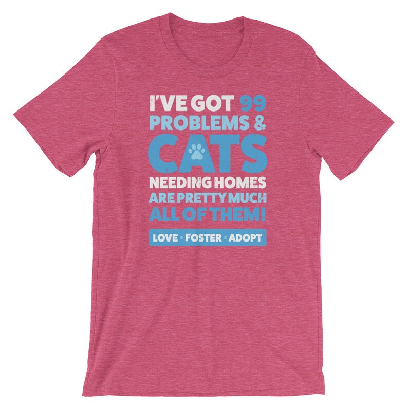 Puede incluir: Una camiseta rosa jaspeada con un gr&aacute;fico azul que dice "Tengo 99 problemas & gatos que necesitan hogar, casi todos! Amar, acoger, adoptar."