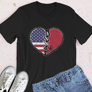 Puede incluir: Camiseta negra con un diseño de corazón roto. El corazón está formado por una bandera estadounidense en un lado y un corazón rojo en el otro. El texto alrededor del corazón dice "Damos nuestro tiempo, nuestras lágrimas, nuestras medallas, nuestras masas, de pie en el camino de la brecha que se ensancha, las heridas profundas y silenciosas de una nación dividida."