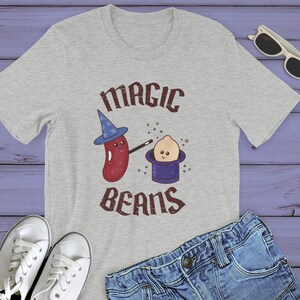 May include: A gray t-shirt with a red bean wearing a wizard hat and holding a magic wand. The bean is pointing the wand at a smaller bean wearing a blue hat. The text "MAGIC BEANS" is printed on the shirt.