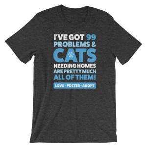Puede incluir: Camiseta gris oscuro jaspeada con una huella de pata azul y el texto "Tengo 99 problemas & gatos que necesitan hogar, son casi todos! Amar, acoger, adoptar."