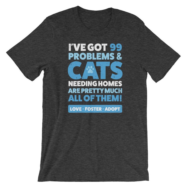 Puede incluir: Camiseta gris oscuro jaspeada con una huella de pata azul y el texto "Tengo 99 problemas & gatos que necesitan hogar, son casi todos! Amar, acoger, adoptar."