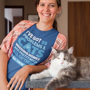 Puede incluir: Una mujer con una camiseta azul con el texto "I've got 99 problems & cats needing homes are pretty much all of them. Love - Foster" est&aacute; sentada con un gato gris y blanco en una encimera de madera.