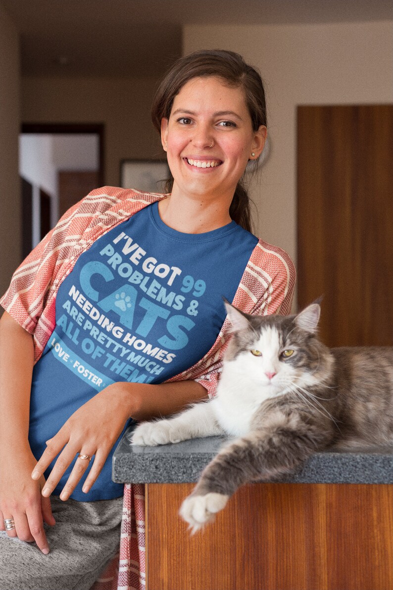 Puede incluir: Una mujer con una camiseta azul con el texto "I've got 99 problems & cats needing homes are pretty much all of them. Love - Foster" est&aacute; sentada con un gato gris y blanco en una encimera de madera.