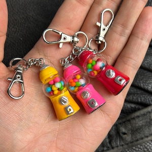 Könnte beinhalten: Drei Schlüsselanhänger in Form von Kaugummiautomaten in Gelb, Pink und Rot. Jeder Schlüsselanhänger hat eine transparente Kugel, die mit kleinen, bunten Kaugummis gefüllt ist. Die Schlüsselanhänger haben silberfarbene Metallverschlüsse und Ketten.