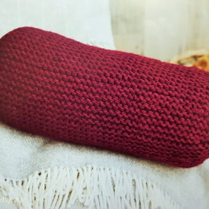 May include: A burgundy-colored, knitted bolster pillow with tassels on each end. The pillow is cylindrical and rests on a white blanket with fringe. The texture of the knit is visible, suggesting a handmade item, perfect for home decor.