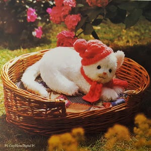 May include: A white plush cat toy wearing a red knit hat and scarf, resting in a woven wicker basket. The cat has black eyes and a pink nose. The basket sits on a patterned fabric.