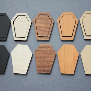May include: Eight wooden coffin-shaped ornaments in various wood finishes, including black, white, brown, and beige. The ornaments are all the same size and shape, and they have a slightly raised edge around the perimeter.
