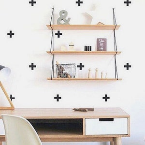 May include: A white wall with black plus signs as wall decor. A wooden shelf with black metal brackets is mounted on the wall. The shelf has three tiers and is decorated with small items, including a book, a small plant, and a wooden figurine. A wooden desk with a white drawer is in the foreground.