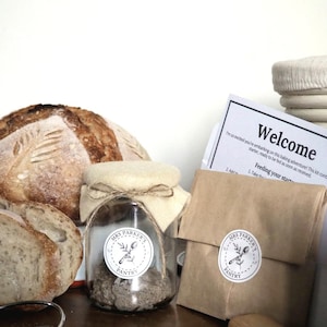 Op de afbeelding: Een glazen pot met een wit deksel en een bruine papieren zak met een wit etiket waarop "Mrs. Parker's Pantry" staat. De pot bevat een zuurdesemstarter. Op de achtergrond is een zuurdesembrood te zien.