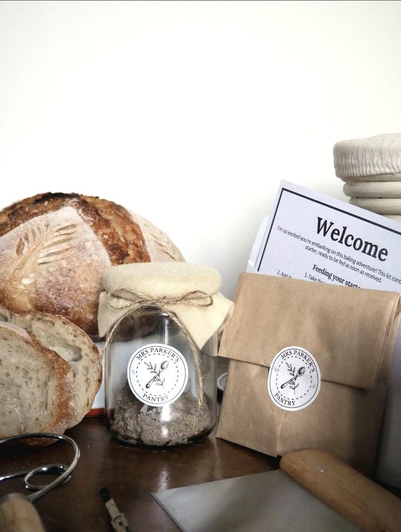 Op de afbeelding: Een glazen pot met een wit deksel en een bruine papieren zak met een wit etiket waarop "Mrs. Parker's Pantry" staat. De pot bevat een zuurdesemstarter. Op de achtergrond is een zuurdesembrood te zien.