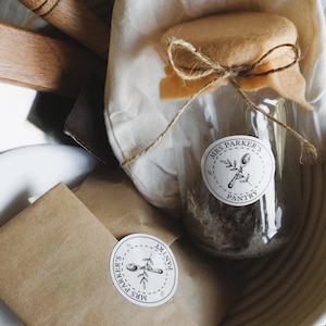 Op de afbeelding: Een close-up van een bruine papieren zak met een witte sticker waarop "Mrs. Parker's Pantry" staat. De zak ligt op een wit oppervlak naast een glazen pot met een bruine deksel en een witte sticker waarop "Mrs. Parker's Pantry" staat.