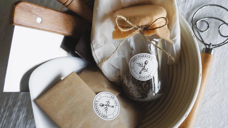 Op de afbeelding: Een close-up van een bruine papieren zak met een witte sticker waarop "Mrs. Parker's Pantry" staat. De zak ligt op een wit oppervlak naast een glazen pot met een bruine deksel en een witte sticker waarop "Mrs. Parker's Pantry" staat.
