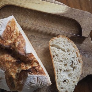 Op de afbeelding: Twee plakken vers gebakken zuurdesembrood op een houten snijplank met een broodmes. Het brood heeft een goudbruine korst en een zacht, luchtig interieur.