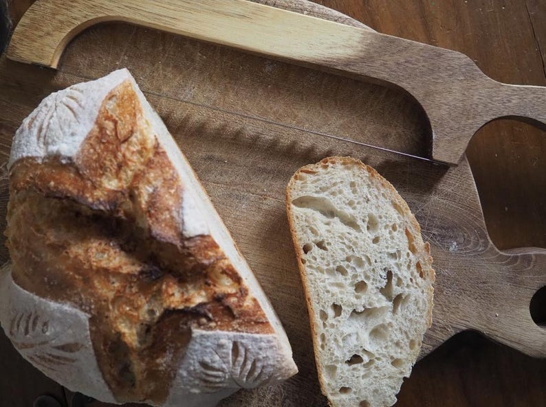 Op de afbeelding: Twee plakken vers gebakken zuurdesembrood op een houten snijplank met een broodmes. Het brood heeft een goudbruine korst en een zacht, luchtig interieur.