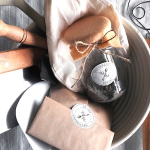 Op de afbeelding: Een witte kom gevuld met bakbenodigdheden, waaronder een glazen pot met een bruin etiket met de tekst "Mrs. Parker's Pantry", een bruine papieren zak met een vergelijkbaar etiket, een houten deegroller, een houten deegkrabber en een witte stoffen zak.