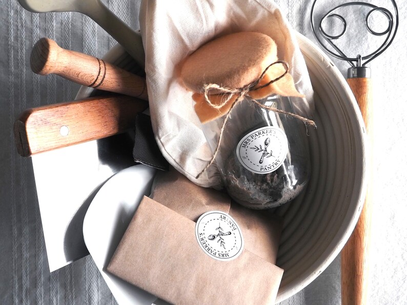 Op de afbeelding: Een witte kom gevuld met bakbenodigdheden, waaronder een glazen pot met een bruin etiket met de tekst "Mrs. Parker's Pantry", een bruine papieren zak met een vergelijkbaar etiket, een houten deegroller, een houten deegkrabber en een witte stoffen zak.