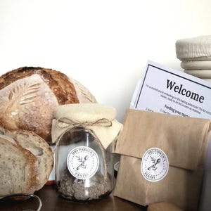 May include: A baking kit with a loaf of sourdough bread, a jar of starter, and a welcome letter. The jar and brown paper bag have "Mrs. Parker's Pantry" labels. The kit includes a wooden rolling pin and other baking tools.
