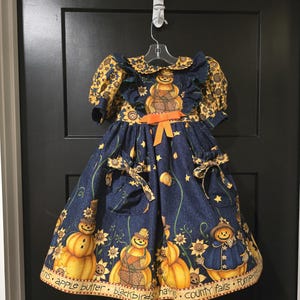 Puede incluir: Vestido azul marino con estampado de calabazas y girasoles. El vestido presenta un cuello y mangas con volantes, un lazo naranja y un diseño de calabaza en el dobladillo. El texto en el dobladillo dice "apple butter, blackbirds, hay, county fairs, pumpkins."