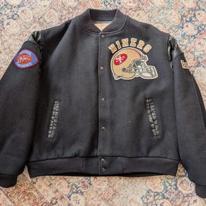 Könnte beinhalten: Eine schwarze Collegejacke mit einem gold-roten Footballhelm-Aufnäher und dem Schriftzug NINERS darüber. Die Jacke hat schwarze Ärmel, einen gerippten Kragen, Bündchen und Saum sowie einen National Football Conference-Aufnäher am rechten Ärmel.