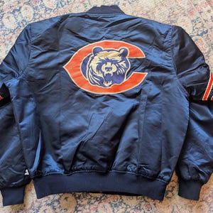 May include: A navy blue satin bomber jacket featuring a large orange and navy bear logo on the back. The sleeves have orange and white striped cuffs, and the jacket includes a ribbed collar and hem, showcasing a classic athletic design.