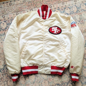 Könnte beinhalten: Eine cremefarbene Satin-Collegejacke mit rot-weiß gestreiftem Kragen, Bündchen und Saum. Auf der Vorderseite befindet sich ein San Francisco 49ers Logo-Aufnäher, während der linke Ärmel ein NFL-Schild-Emblem und ein kleines Starter-Logo zeigt.