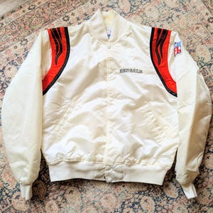 May include: A white satin bomber jacket featuring the word BENGALS embroidered on the chest. The jacket has red and black flame-like accents on the shoulders, a snap-button front closure, and an NFL logo patch on the upper left sleeve.