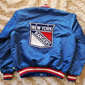 May include: A blue satin jacket featuring the New York Rangers logo. The jacket has red, white, and blue detailing on the collar and cuffs. A vintage style jacket.