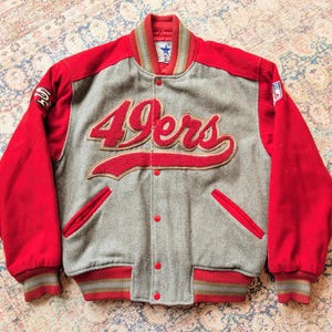May include: A red and grey varsity jacket featuring the text 49ers in red felt lettering across the chest. The jacket has red sleeves, a grey torso, a ribbed collar and cuffs with striped detailing, red snap buttons, and two side pockets.