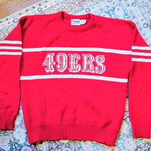 Könnte beinhalten: Ein leuchtend roter Langarmpullover mit Rundhalsausschnitt und weißen horizontalen Streifen auf Brust und Ärmeln. Das Wort 49ERS ist in weißen Blockbuchstaben mittig auf die Brust gestrickt. Das Kleidungsstück hat gerippte Bündchen am Kragen, Ärmeln und Saum.