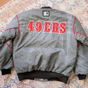 May include: A grey Starter brand jacket featuring a pinstripe pattern and red piping. The back displays the text 49ERS in red lettering with a white outline. The jacket includes black ribbed cuffs and a black ribbed hem with a snap button closure.