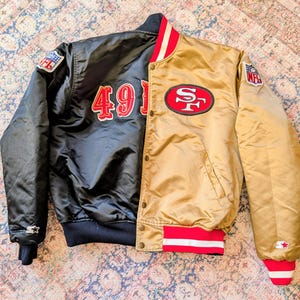 Könnte beinhalten: Eine zweifarbige Collegejacke mit einer schwarzen Seite mit dem rot-weißen Schriftzug 49ers und einer goldenen Seite mit einem rot-weißen 49ers-Logo. Die Jacke verfügt über NFL-Aufnäher an den Ärmeln, eine Druckknopfleiste und rot-weiß gestreifte Bündchen.