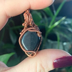 Peut inclure: Un pendentif en fil de cuivre enroulé autour d'une pierre précieuse gris foncé. Le pendentif est tenu dans une main avec un vernis à ongles rouge foncé.