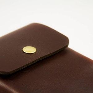 May include: Close-up of a dark brown leather case with a flap and a gold-colored button closure. The leather has a smooth texture and the case appears to be well-crafted. The background is plain white.