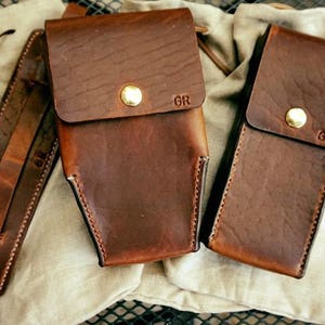 May include: A collection of handcrafted brown leather goods, including two rectangular cases with flap closures and a shoulder strap. Each case features a gold-colored button and the initials "GR". The items are displayed on a neutral-colored fabric.