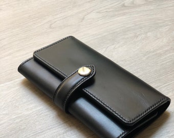 May include: A black leather wallet with a gold button closure and a strap. The wallet is folded in half and lying on a light wood surface.
