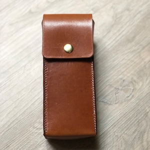 May include: A brown leather case with a brass button closure. The case has a stitched edge and is designed to hold a small object.