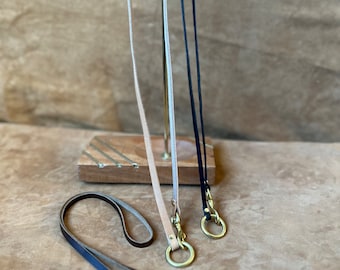 May include: Three leather keychains with brass hardware. The keychains are hanging on a metal display stand. The keychains are brown, beige, and black.