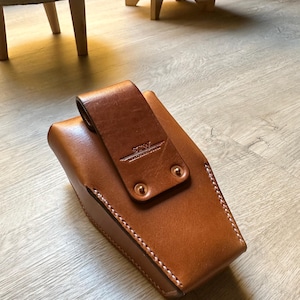 May include: A brown leather case with a flap closure and white stitching. The case has a belt loop and is secured with two copper rivets. The leather has a smooth texture and a natural color.