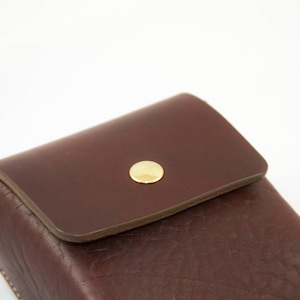 May include: A dark brown leather case with a flap closure and a gold-colored button. The case has a textured surface and appears to be handmade. The design is simple and classic.