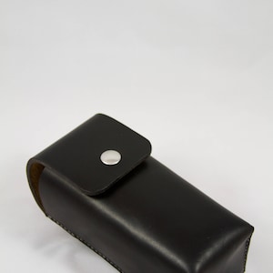 May include: A black leather glasses case with a silver press stud closure. The rectangular case has rounded edges and a flap secured by a single, circular silver press stud. The case is designed to protect spectacles.