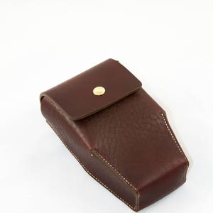 May include: A rectangular, dark brown leather case with a flap closure and a gold-colored button. The case is stitched with white thread and appears to be handmade. The leather has a textured appearance.