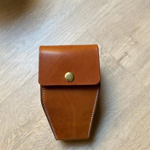 May include: A brown leather case with a flap secured by a gold-colored button. The case has a trapezoidal shape and white stitching along the edges. The leather has a smooth texture.